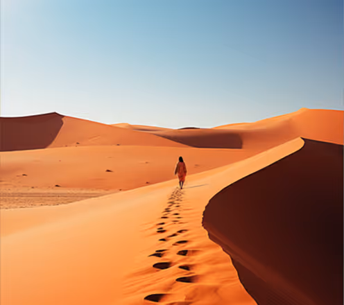 Person walking alone on a ridge of large sand dunes under clear blue sky.