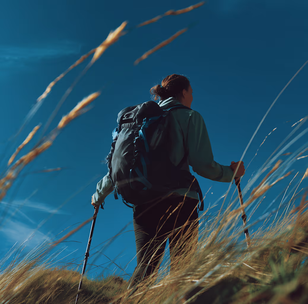 Hiker with backpack walking with trekking poles through tall grass under clear blue sky.