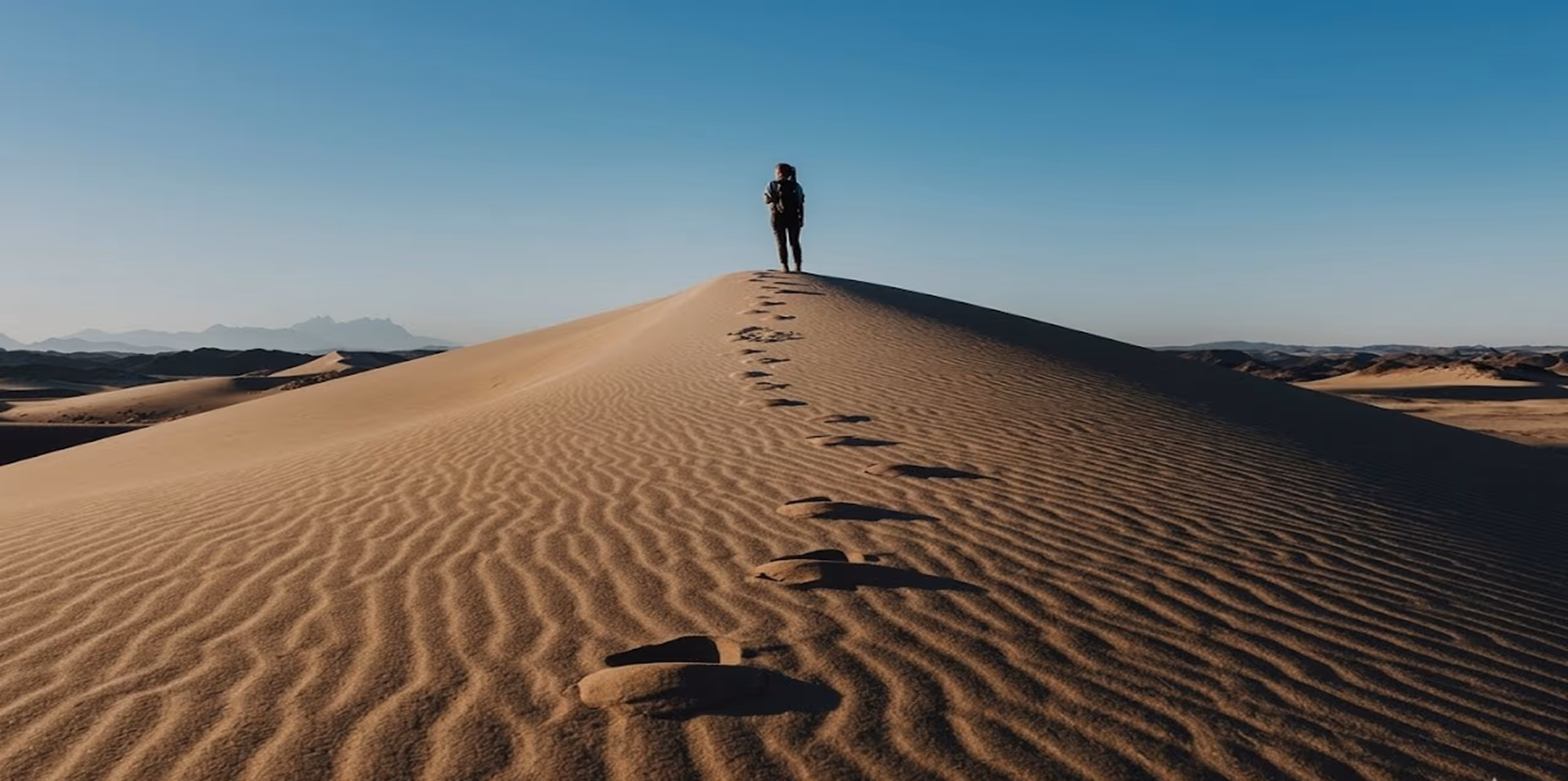 Person standing on top of a sand dune with footprints leading up under clear blue sky.