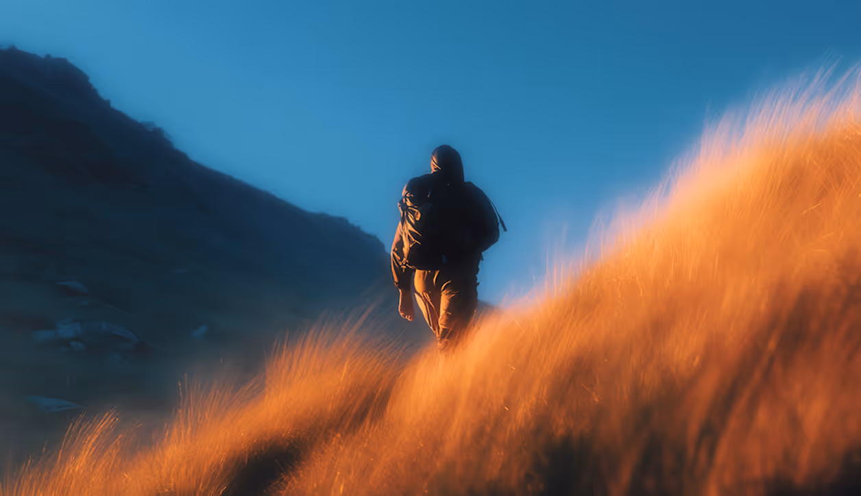 A hiker with a backpack walking up a grassy hill at sunset with a blue sky background.