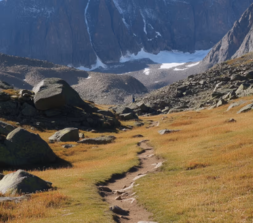 Narrow dirt trail through rocky alpine meadow leading toward snow patches below steep mountain cliffs.