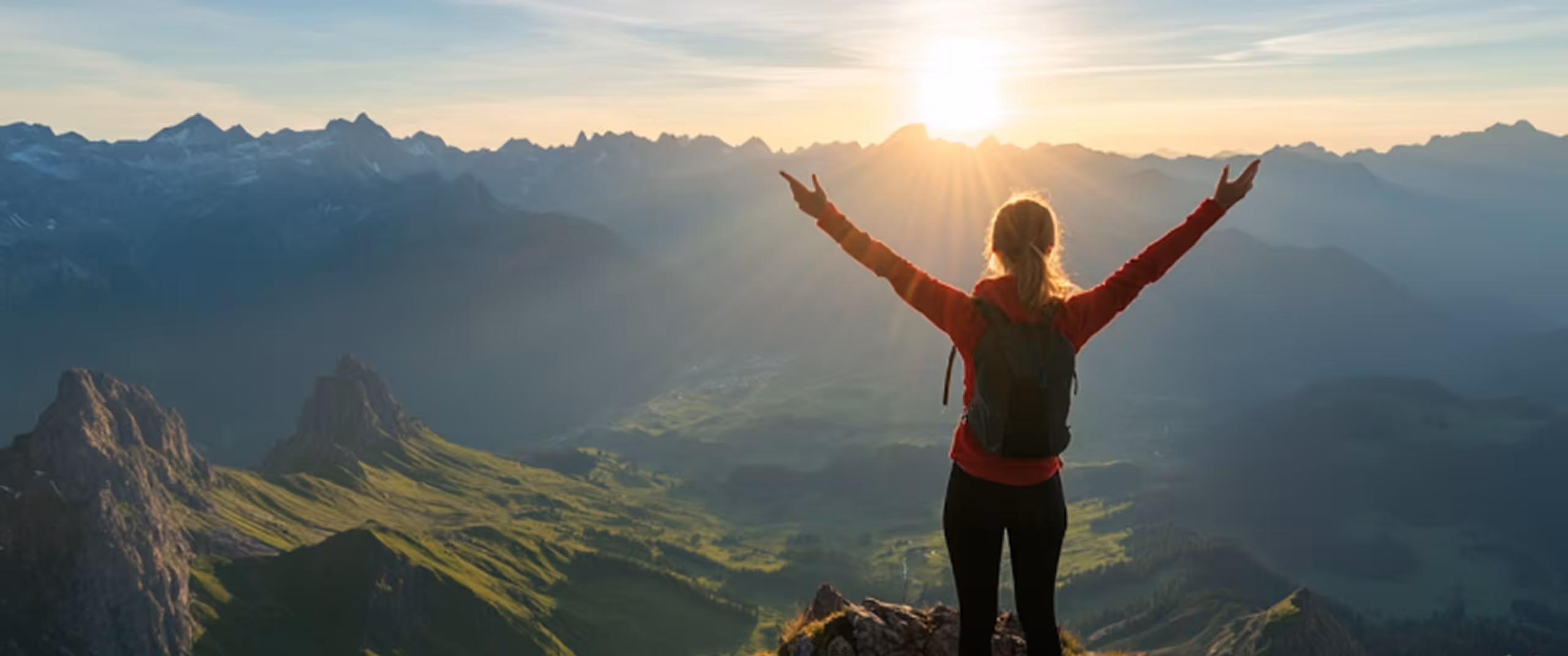 Hiker with arms raised overlooking a sunlit mountain valley at sunrise or sunset.