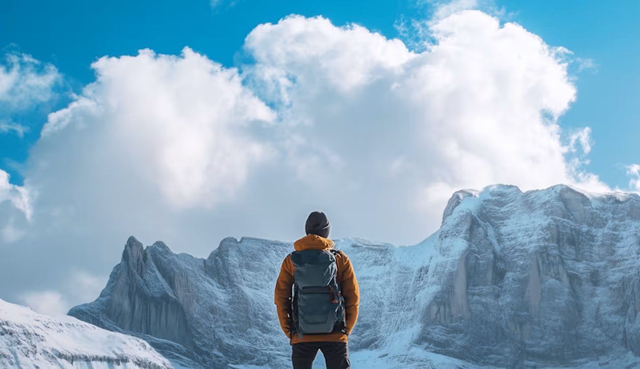Back view of a person in orange jacket with backpack facing snowy mountain under blue sky with clouds.