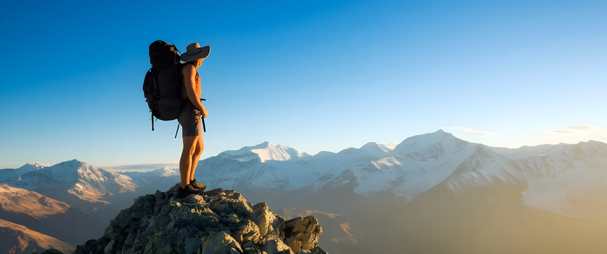Hiker with backpack and hat stands on rocky peak overlooking snow-capped mountains at sunrise.