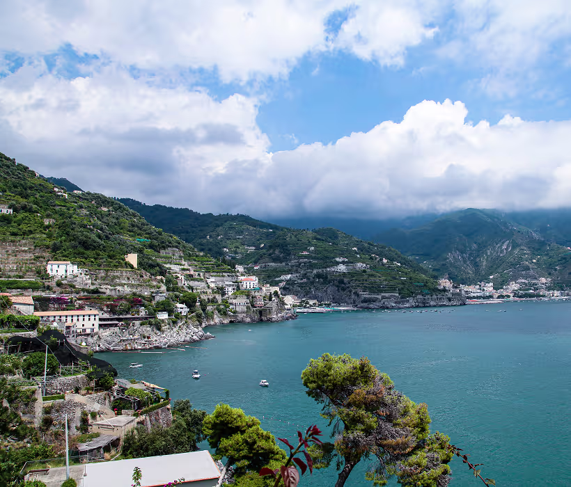 Coastal landscape with terraced hillsides, scattered houses, turquoise sea, and cloudy sky.