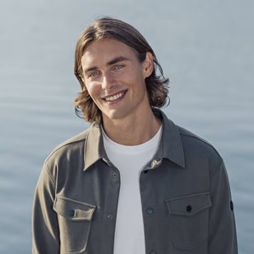 Young man with shoulder-length hair smiling, wearing a gray jacket over a white shirt by water.