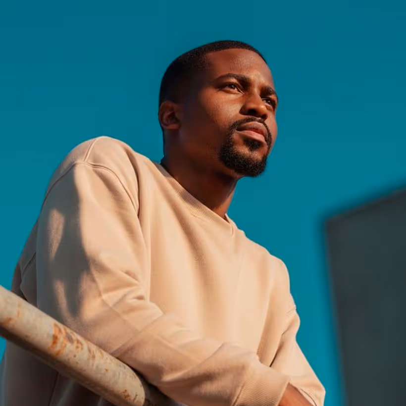 Man with a beard wearing a beige sweatshirt looking thoughtfully into the distance against a blue sky.
