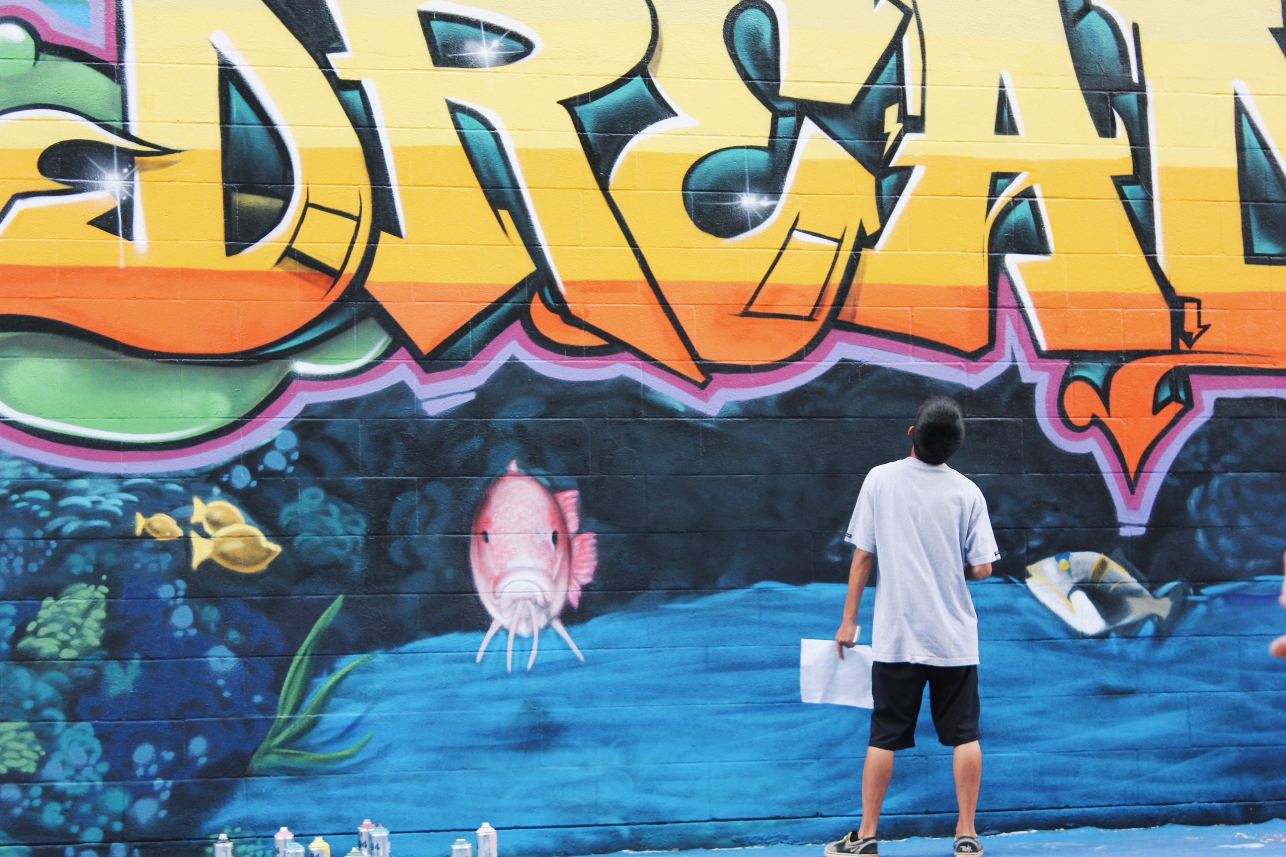 Trysen, then a Kauaʻi High senior, looking up at the Dreams lettering with his sketch in hand and spray cans at his feet