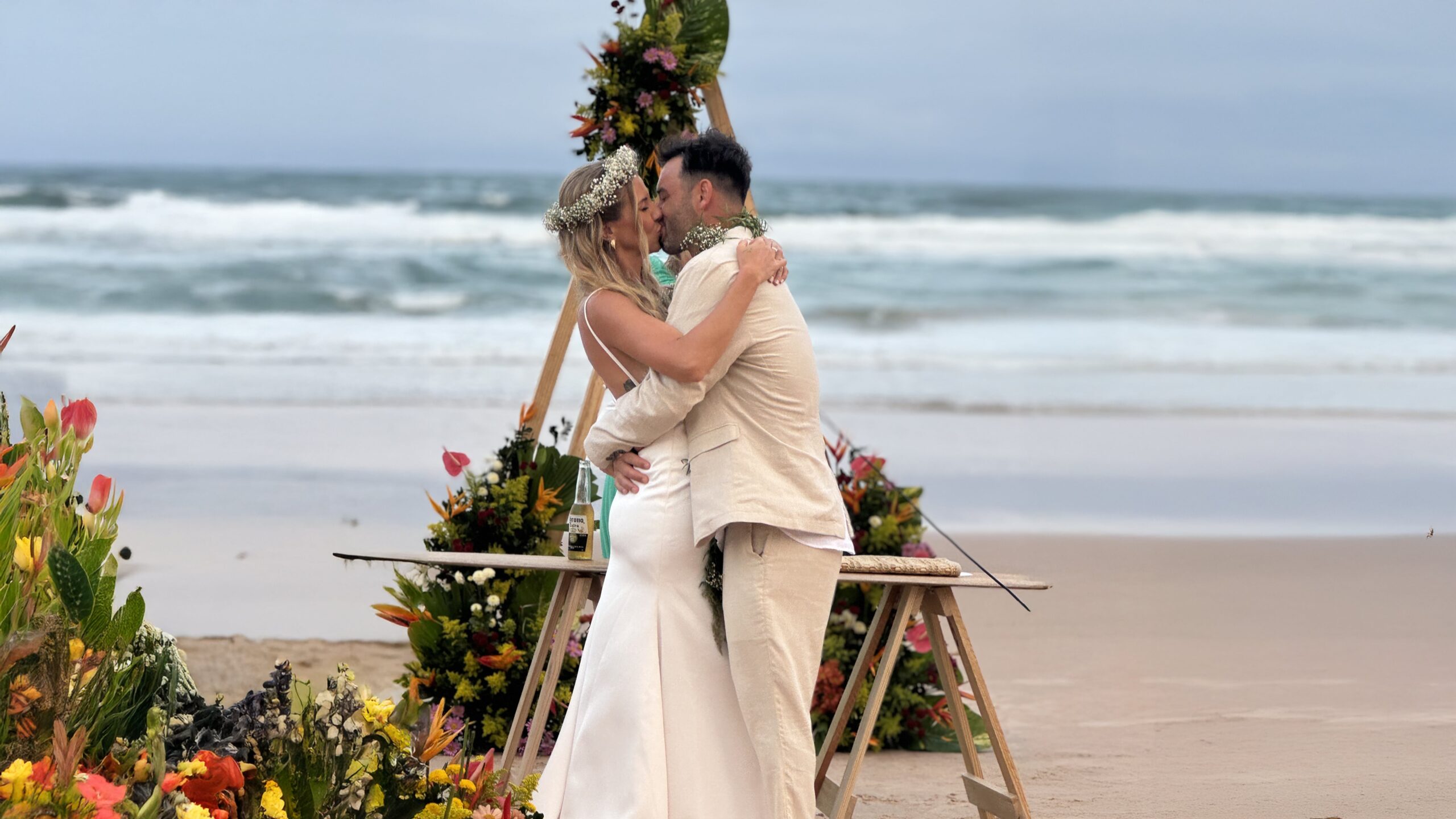 Casal beijando em casamento na praia com flores coloridas e mar ao fundo.