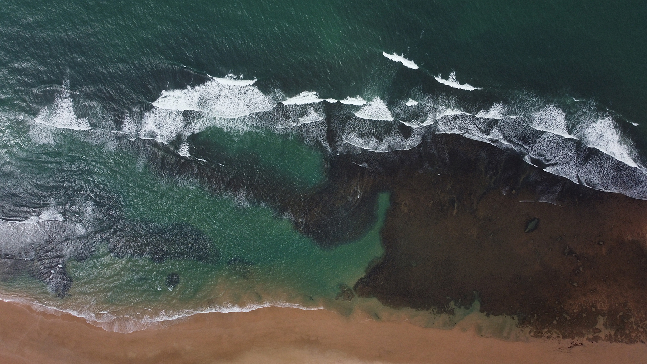 Vista aérea da praia com ondas quebrando na costa e areia marrom.