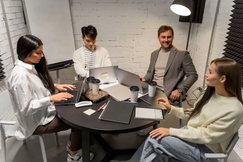 Four diverse colleagues at a round table in a modern office, working on laptops and discussing.