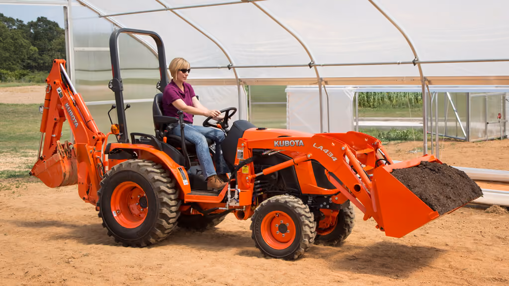 Operator driving an orange Kubota compact tractor with loader and backhoe near a greenhouse.
