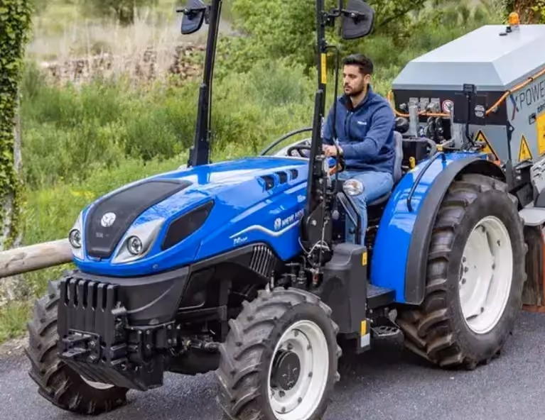 Operator driving a blue New Holland T4 series tractor along a farm track with a mounted implement behind.