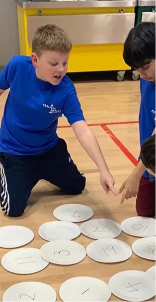 Two boys in blue shirts kneeling on floor, pointing at paper plates with numbers written on them.