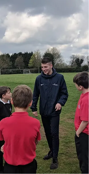 Young coach talking to four boys on a grassy sports field with cloudy sky overhead.