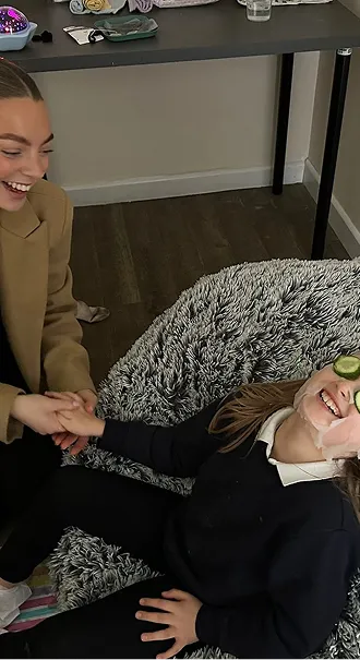 Woman holding hands with smiling girl lying on a fluffy beanbag with cucumber slices on her eyes.