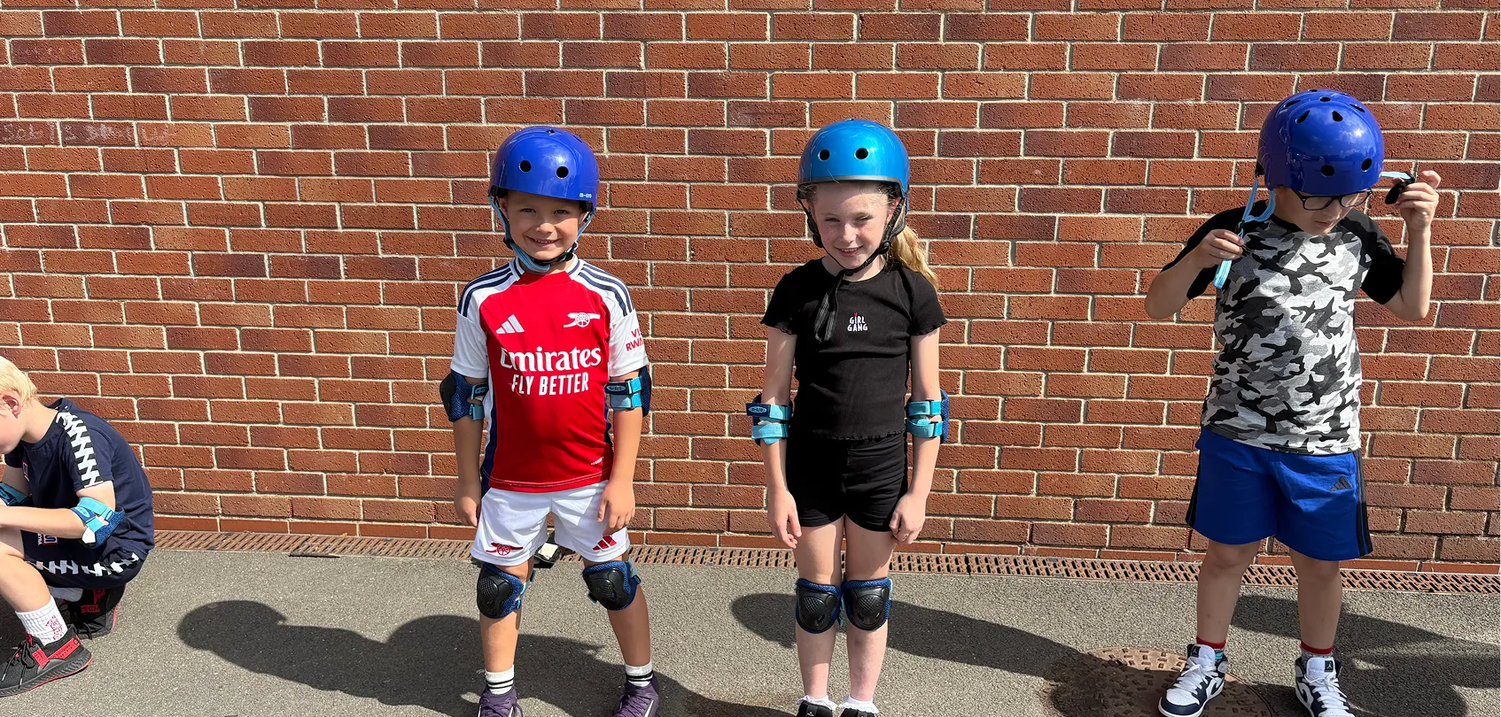 Three children wearing helmets and knee pads stand against a brick wall on a sunny day.