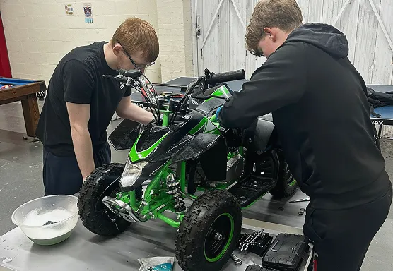 Two boys working on a green and black ATV indoors on a table with tools and a bowl nearby.