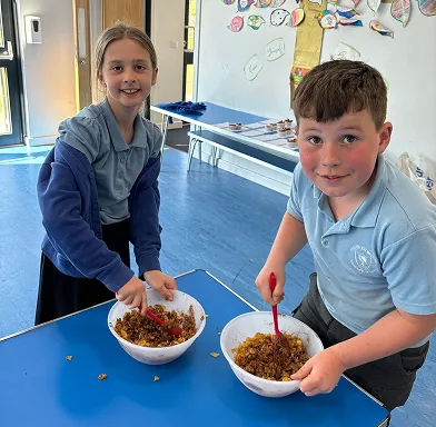 Two children mixing food ingredients in white bowls on a blue table in a classroom.
