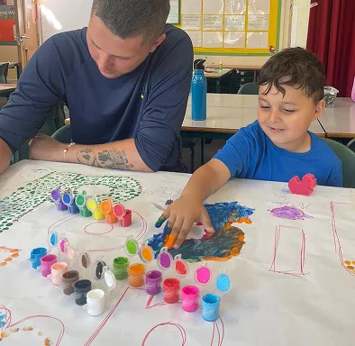 Child finger painting on paper while adult watches at a classroom table with colorful paints.
