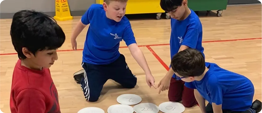 Four boys, three wearing blue shirts, kneeling on gym floor pointing at numbered paper plates.