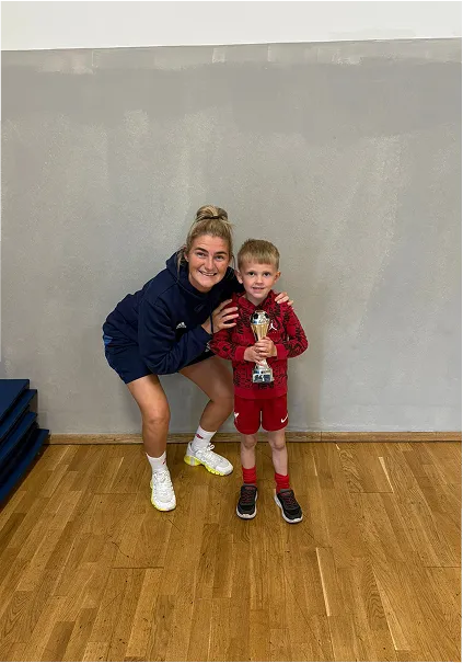 Woman in sportswear crouching next to a young boy holding a trophy on a wooden floor.