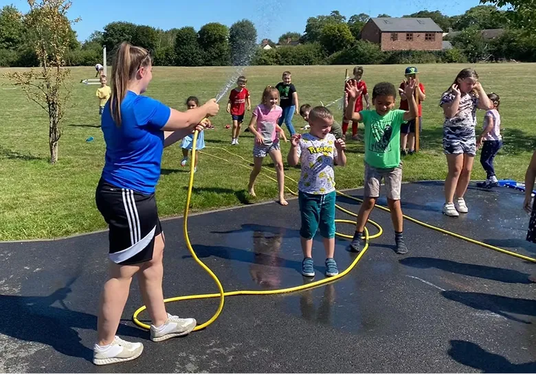 Woman spraying children with a hose as they play and jump on wet pavement outdoors.
