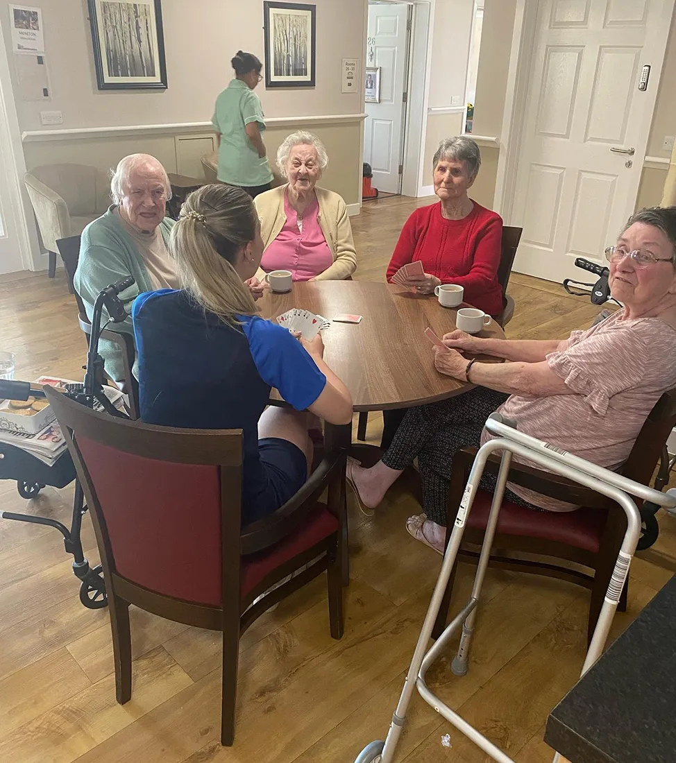 Four elderly women and one caregiver sitting around a table playing cards and drinking tea in a care home.