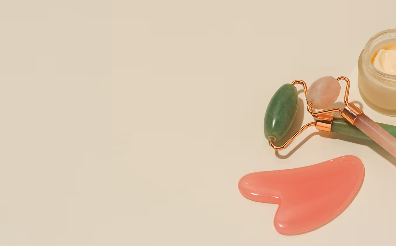 Two jade facial rollers, a pink gua sha tool, and a jar of cream on a beige surface.