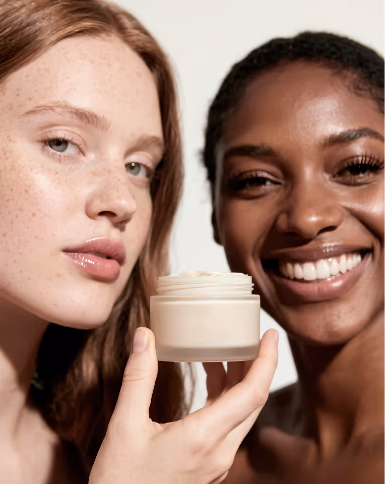 Two women, one holding a jar of cream, smiling and showcasing skincare product close-up.