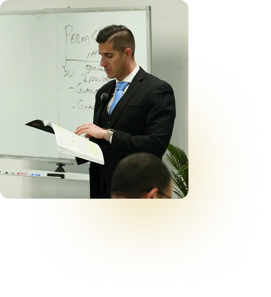 Man in suit reading from a book in front of a whiteboard with writing in a classroom setting.