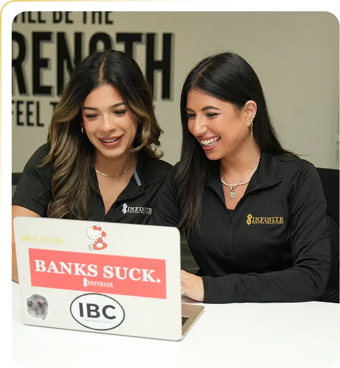 Two women in black Infinite Capital Solutions shirts smiling while looking at a laptop with stickers.