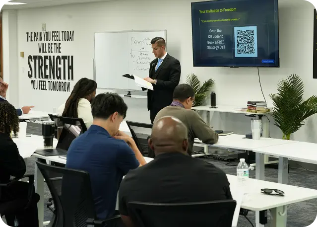 A man in suit leads a class with seated attendees, motivational quote on wall, and QR code on screen.
