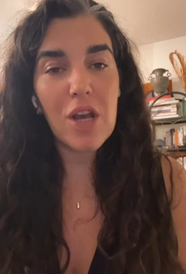 Woman with long curly dark hair speaking in a room with shelves and books behind her.