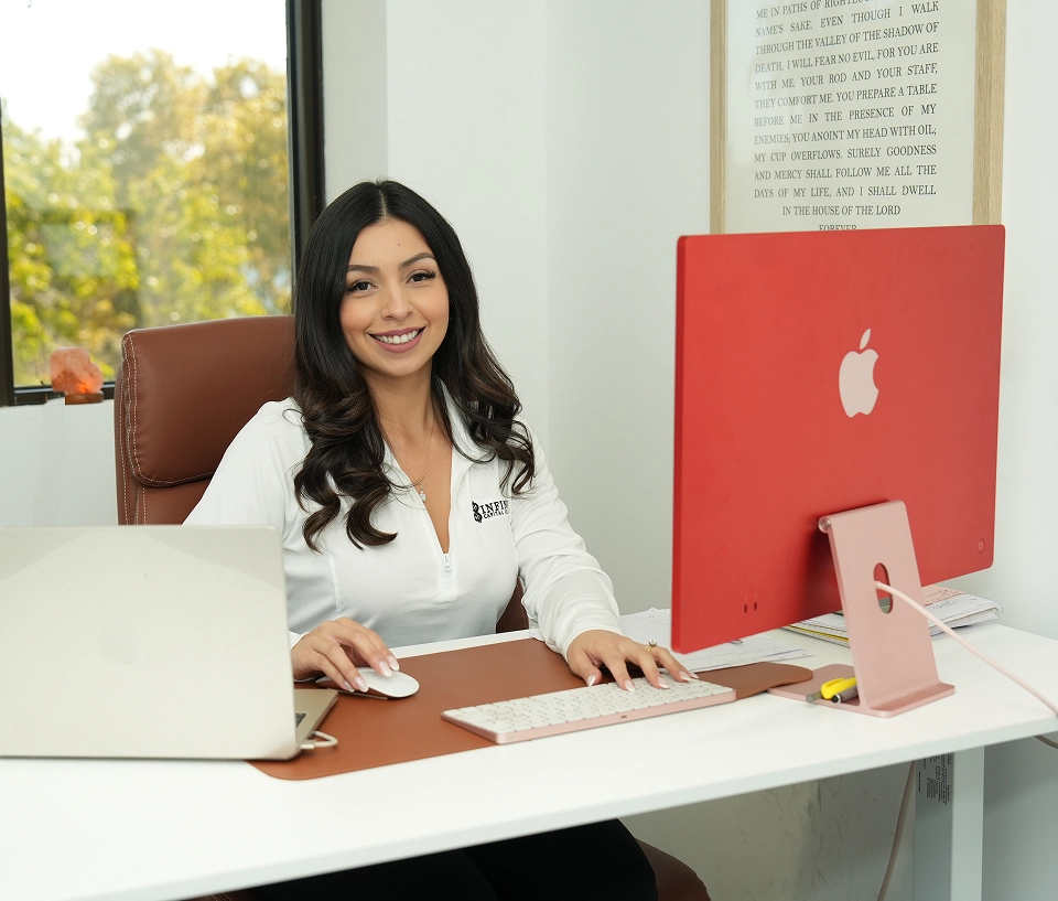 Smiling woman with long dark hair sitting at desk with a red iMac and laptop in bright office.