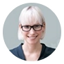 Smiling woman with glasses and light hair wearing a black top, against a pale blue background.