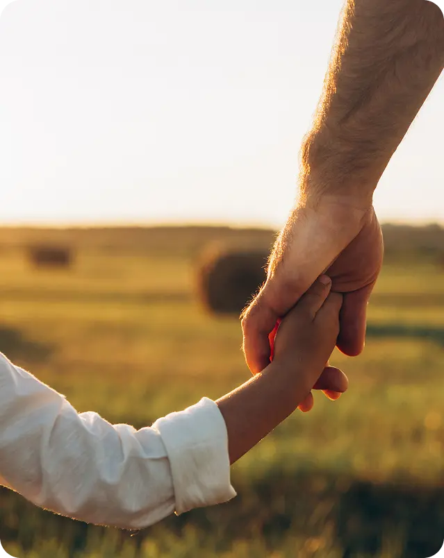 Adult and child holding hands outdoors in a sunlit field with blurred hay bales in the background.