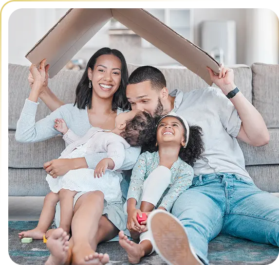 Smiling family of four sitting on floor, parents holding cardboard roof over their heads in living room.