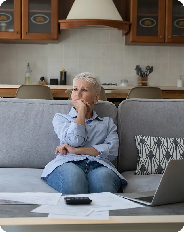 Pensive elderly woman sitting on a couch with papers and laptop on the table in front of her.