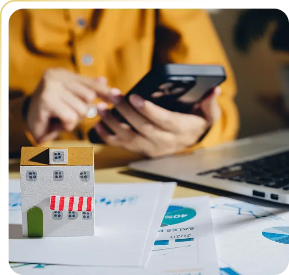 Person using smartphone near laptop and miniature house model on financial papers.