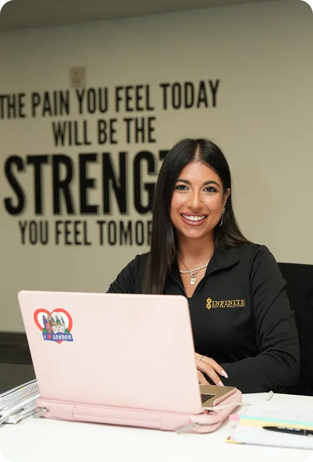 Smiling woman with dark hair sits at a desk with a laptop, motivational quote on wall behind her.