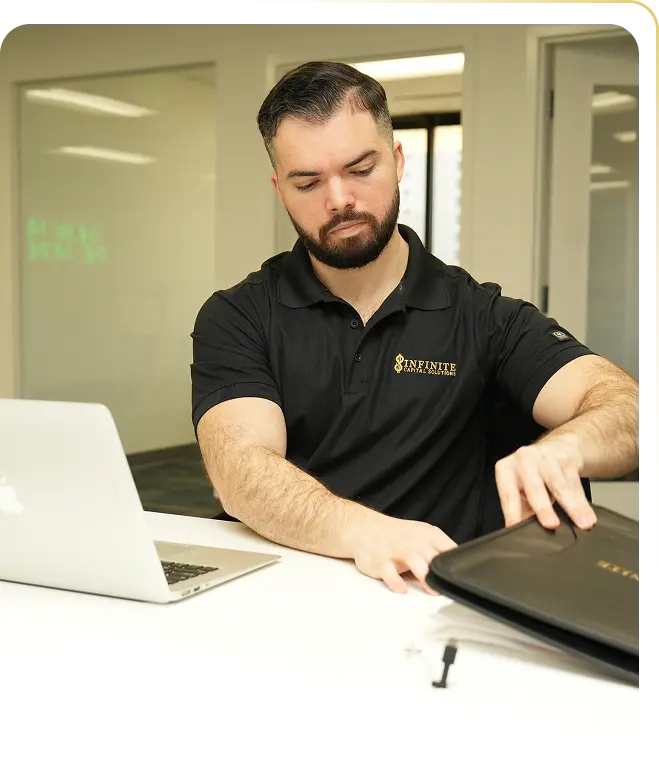 Man with beard in black Infinite Capital Solutions polo organizes documents at desk with laptop.