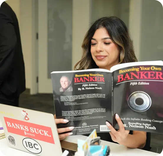 Woman sitting at desk reading the book Becoming Your Own Banker by R. Nelson Nash.