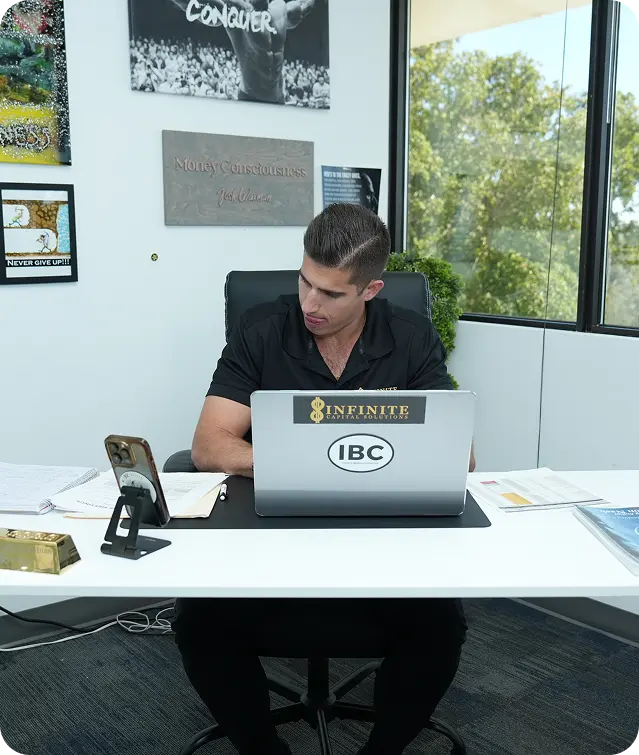 Man in black shirt working at a desk with laptop, phone, papers, and motivational posters on the wall.