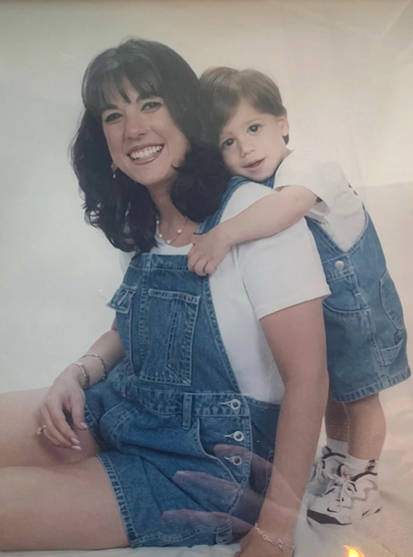 Smiling woman in denim overalls with child hugging her from behind, both wearing white shirts and denim.