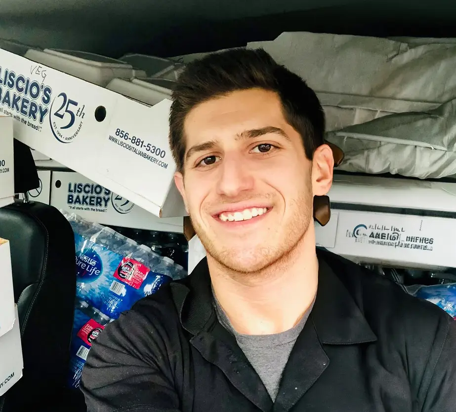 Smiling young man wearing black shirt inside a vehicle loaded with bakery boxes and water bottles.