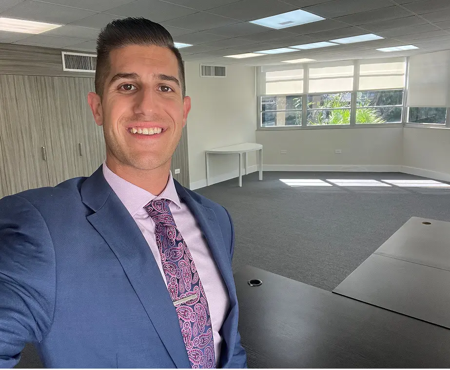 Smiling man in a blue suit and patterned tie taking a selfie inside a bright, empty office room.