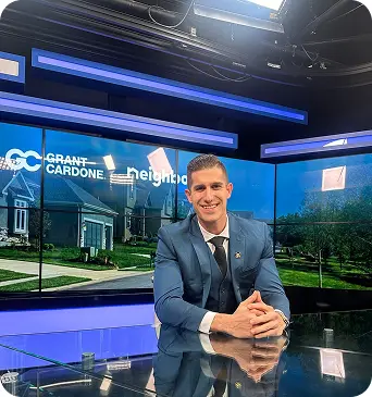 Man in navy blue suit sitting at reflective desk in TV studio with Grant Cardone branding behind him.