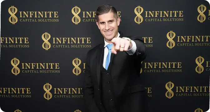 Man in suit smiling and pointing towards the camera in front of Infinite Capital Solutions backdrop.