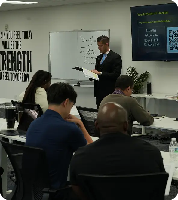 Man in suit presenting to seated audience in a classroom with whiteboard and motivational quote on wall.